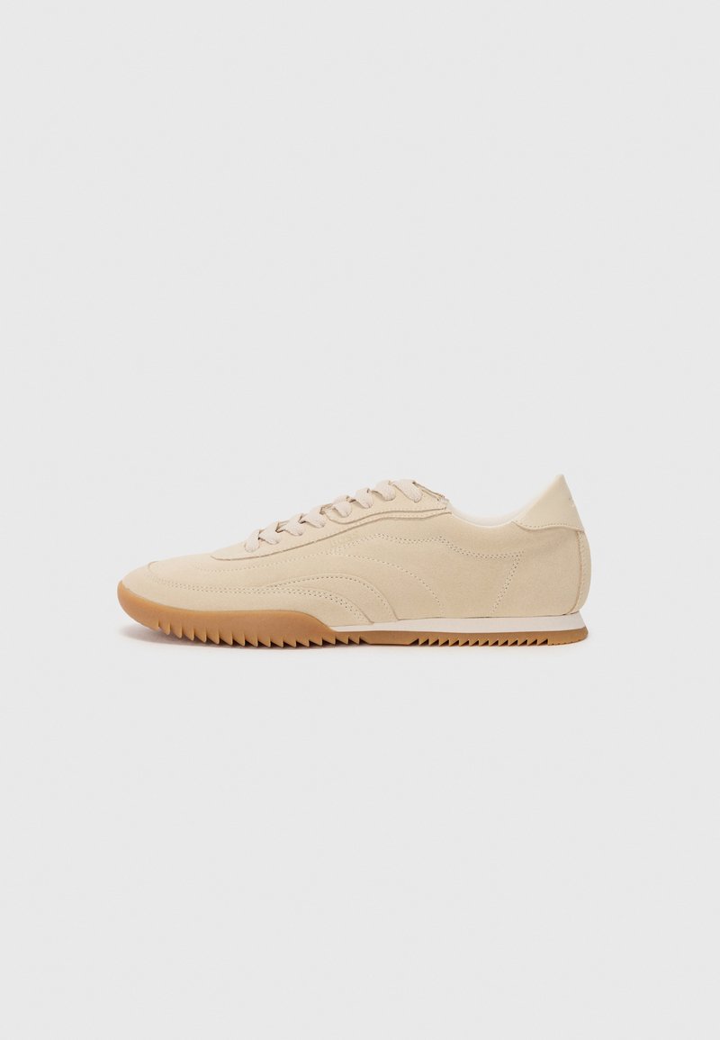 Beige ruskinn sneakers med et strømlinjeformet design, med en teksturert overdel, rund tå, og en gummisåle med et sikksakk-mønster.