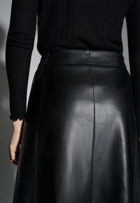 Jupe en cuir noir à texture lisse, présentant des coutures visibles et une taille ajustée. Haut noir côtelé partiellement visible.