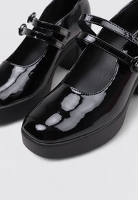 Zapatos Mary Jane de charol negro con dos correas delgadas, punta redondeada y suela de plataforma gruesa. Acabado suave y brillante con sutiles reflejos.
