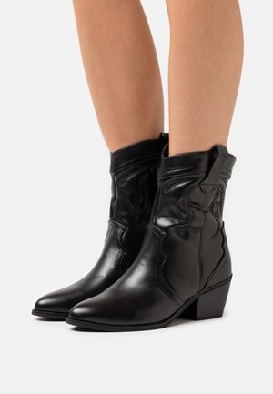 Botas de vaquero negras de cuero hasta el tobillo con detalles de costura en forma de llama, usadas sobre piernas desnudas, con tacones apilados y pestañas de agarre.