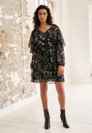 Schwarzes Blumenkleid mit durchscheinendem Überzug, V-Ausschnitt und geraffter Taille. Fließende Ärmel und strukturierte Knöchelstiefel runden den Look ab.