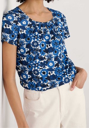 Kurzärmeliges blau-weißes florales Oberteil mit plissiertem Halsausschnitt, kombiniert mit hoch taillierter cremefarbener Hose, Hand in der Tasche.