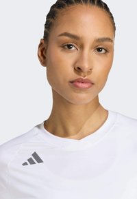 Bijela atletska majica s V-izrezom, izrađena od glatke tkanine, sivosivi Adidas logo na lijevom ramenu.