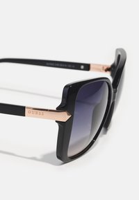 Ochelari de soare negri cu o ramă groasă, lentile gri închis cu gradient și accente roz aurii pe tâmple, având logo-ul "GUESS."