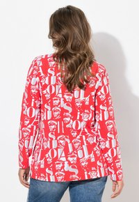 Rotes Langarmshirt mit einem auffälligen, sich wiederholenden Muster aus weißem Text und illustrierten Gesichtern, kombiniert mit blauen Jeans.