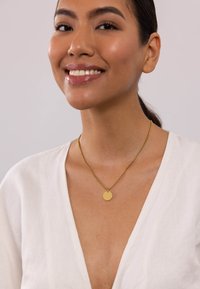 Goldene Anhängerhalskette an einer feinen Kette mit einer runden Scheibe als Blickfang. Das Model trägt ein weißes V-Ausschnitt-Top. Glatte Textur, schlichtes Design.