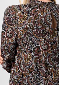 Schwarze Stoffbluse mit filigranem Paisleymuster in Rot, Grün und Weiß. Verfügt über einen gerafften Ausschnitt und eine Schlüssellochöffnung am Rücken.