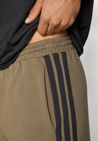 Olivzaļas sporta bikses ar ribotu jostas vietu, tām ir trīs tumšas svītras pa sānu un sānu kabata. Mīksts, elastīgs audums.