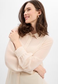Kremowa, przezroczysta bluzka z zapięciem na guziki i kołnierzykiem. Posiada bufiaste rękawy z elastycznymi mankietami oraz subtelne haftowane logo marki na rękawie.