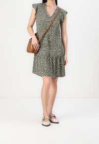 Kleid mit Blumenmuster in Dunkelgrün, mit V-Ausschnitt, Rüschensleeves und gestuftem Saum. Kombiniert mit hellen flachen Schuhen und einer braunen Schultertasche.