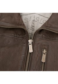 Braune Lederjacke mit Reißverschluss, strukturiertem Oberflächen und metallischen Reißverschlussanhängern mit Markenname. Innenfutter hat eine helle Auskleidung.