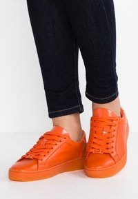 Oranžové nízké tenisky vyrobené z lesklého materiálu mají ploché tkaničky, zaoblenou špičku a mírně texturovanou podrážku. Nosené s tmavými džíny.