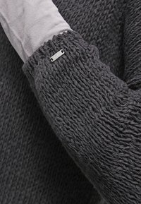Maglione lavorato a maglia spesso con una texture grigio scuro; presenta una manica interna di un grigio chiaro a contrasto e un piccolo logo metallico sul polsino.