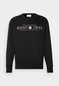 Svart hettegenser laget av bomull, med rund hals, lange ermer, og et grafisk trykk med "GANT 1949" og et skjoldemblem foran.