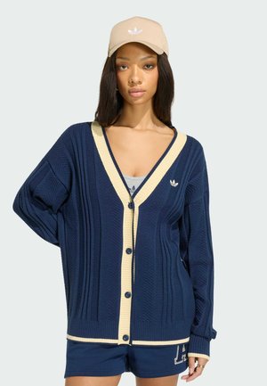 Junge Frau, die eine beige Kappe, eine marineblaue Strickjacke mit cremefarbenem Saum, ein graues Tanktop und marineblaue Shorts trägt, steht mit einer Hand in der Hüfte.
