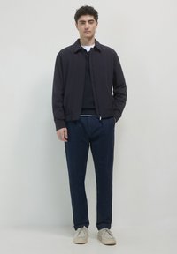 Giubbotto bomber blu navy con colletto, chiusura a zip e polsini elasticizzati, abbinato a pantaloni in denim scuri e scarpe di colore chiaro.