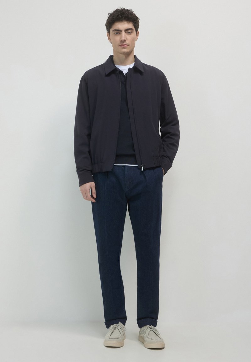 Giubbotto bomber blu navy con colletto, chiusura a zip e polsini elasticizzati, abbinato a pantaloni in denim scuri e scarpe di colore chiaro.