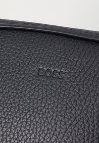 Geantă din piele neagră cu o suprafață texturată, logo-ul „BOSS” embosat și un margine netedă, cosută. Formă rectangulară compactă.