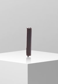 Un dispositif rectangulaire marron avec une texture lisse, présentant une extrémité arrondie et un profil fin, reposant verticalement sur un piédestal blanc.