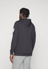 Schwarzer Hoodie mit Kängurutasche, gerippten Bündchen und Saum. Verfügt über ein Logo-Patch am linken Ärmel. Getragen mit grauen Sweatpants.