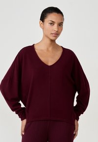 Kobieta z ciemnymi włosami związanymi w kok, ubrana w luźny, ciemno-burgundowy sweter z dekoltem w serek i pasujące prążkowane spodnie, stojąca z rękami w kieszeniach.