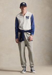Férfi tengerészkék és fehér sapkában, fehér és tengerészkék baseballingben logóval, szürke tréningnadrágban, derekán megkötött tengerészkék pulóverrel és tengerészkék sportcipőben.