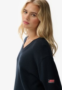 Blonde vrouw met losse golven draagt een marineblauwe V-halstrui met een kleine Amerikaanse vlagpatch op de linkermouw, terwijl ze over haar schouder kijkt.