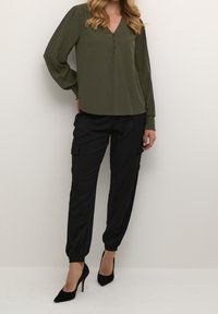 Blouse kaki à manches longues avec col en V et détails de boutons, associée à un pantalon cargo noir avec poignets élastiques et poches, portée avec des talons noirs.