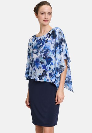 Hellbluse mit floralem Muster und fließenden Umhangärmeln, die über einem figurnahen blauen Rock getragen wird. Leichtes Material mit glatter Textur und lebendiger Farbe.