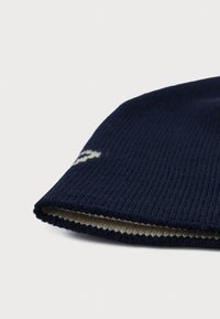 Strikket marineblå lue med ribbestruktur, med en kremfarget fôr og en liten logodetalj på siden.