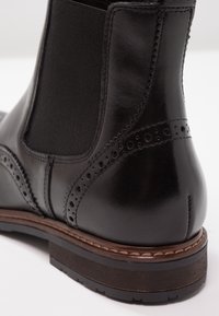 Botín Chelsea de cuero negro con paneles laterales elásticos, detalles de brogue decorativos y una suela marrón robusta con costuras.