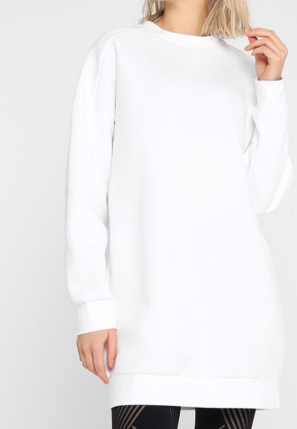 Personne portant un sweat-shirt blanc long et ample avec des poignets et un ourlet en côte, associé à des leggings noirs à motifs sur un fond uni.