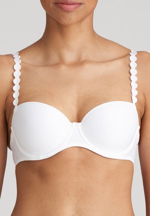 Vrouw draagt een witte gewatteerde beha met hartvormige sierbandjes, toont schouders en bovenborst tegen een neutrale achtergrond.