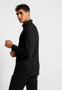 Melnā krekls, kas izgatavots no gludas auduma, ar pogu kaklu, garām piedurknēm un nedaudz noapaļotu apakšmalu. Slim-fit dizains.