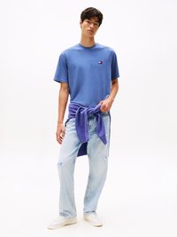 T-shirt in cotone blu con un piccolo logo sul petto, abbinata a jeans chiari strappati. Un maglione viola è annodato in vita.