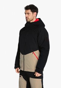 Schwarze und beige Jacke mit Kapuze, die ein diagonales Design aufweist, rote Reißverschlussakzente und strukturierten Stoff hat.