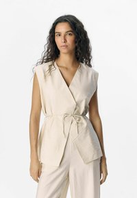 Top avvolgente senza maniche beige con cintura da annodare, caratterizzato da scollatura a V e tessuto testurizzato. Design semplice senza ulteriori motivi o accenti.