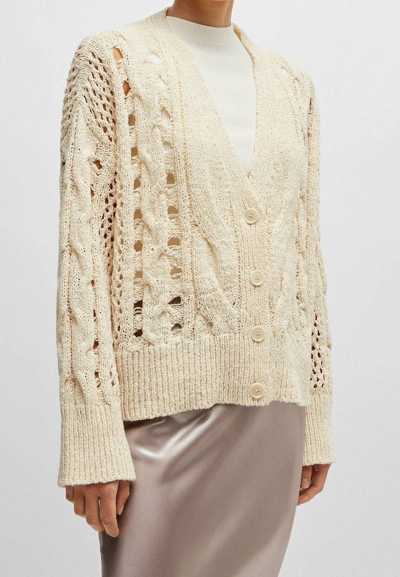 Kvinna iklädd en krämfärgad kabelstickad cardigan med knappar över en vit polotröja och en glänsande beige kjol, visad från halsen till höfterna.