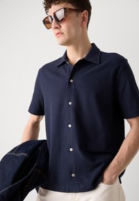 Camicia a maniche corte blu navy realizzata in un tessuto texturizzato, con chiusura a bottoni, colletto a punta e bottoni coordinati. Indossata con occhiali da sole.
