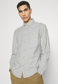 Camisa de manga larga gris claro hecha de una mezcla de algodón, con cuello abotonado, bolsillo en el pecho y botones beige, sobre un fondo liso.