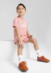 Rožinė medvilninė marškinėlė su balta "PUMA" logotipu, atitinkamos rožinės šortai, oranžiniai ir purpuriniai sportbačiai bei balti kojinės su juodu logotipu. Sėdima poza.