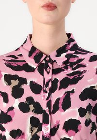 Košile na knoflíky z růžové látky s černo-hnědým leopardím potiskem. Má klasický límec a růžové knoflíky podél přední části.