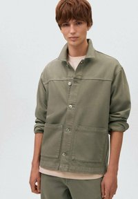 Olivnozelena jeans jakna s ovratnikom, gumbom na sprednji strani, dvema sprednjima žepoma in zavihanimi rokavi, nosi se čez svetlo srajco.