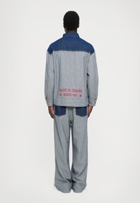 Personne de dos portant une veste en denim grise et bleue ainsi qu'un pantalon, avec le texte rouge "FAIT AU CIEL DEPUIS 1967" dans le dos de la veste.