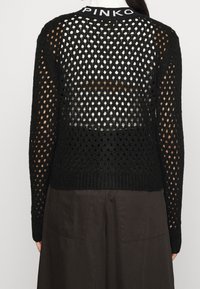 Svart stickad tröja med löst, öppet mönster, utrustad med ribbad fåll och en märkesdetalj vid halsen. Baksidan är till största delen genomskinlig.