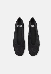 Un paio di scarpe nere slip-on con ricami floreali, punta quadrata e etichette "Camper" e "Twins" all'interno, mostrate dall'alto su uno sfondo bianco.