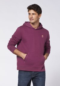 Purple cotton hoodie with a front pocket, ribbed cuffs, and an embroidered logo on the chest. Worn with dark denim jeans.