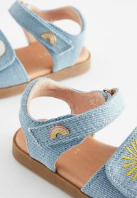 Blå denimsandaler med två justerbara remmar, tanfärgade innersulor och en regnbågsdetalj på remmen. Har gul broderi på framsidan.