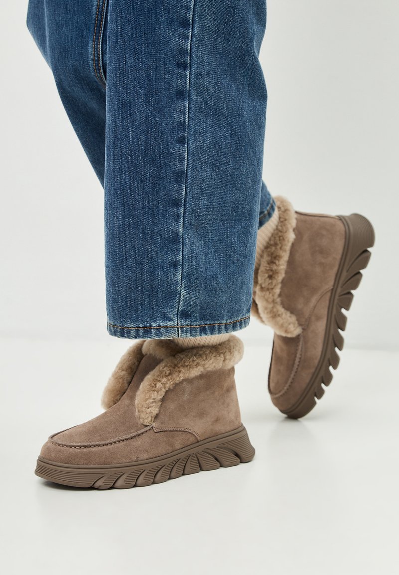 Pies que lucen botas de invierno de ante beige con forro de piel de oveja y suelas gruesas, combinadas con jeans de mezclilla azules y calcetas beige.