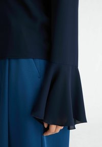 Námořnická blůza se zvonovými průhlednými rukávy, spárovaná s modrými kalhotami. Hladká textura látky, elegantní design. Žádné viditelné vzory ani ozdobné prvky.